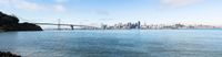 city skyline, bridge and ocean with rocks on shore against clear blue sky, san francisco california bay