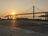 two men with skateboards stand in front of the water near the bridge as the sun sets