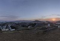 the view from top of a hill at sunset, overlooking a small city below mountains