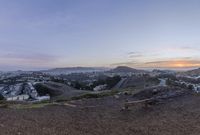 the view from top of a hill at sunset, overlooking a small city below mountains