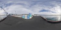 fish eye view of street with parking area and boat ramp by water, under a cloudy sky