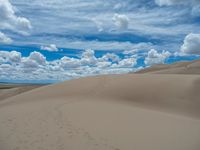 a lone person walks through a desert like area surrounded by sand dunes and bright blue sky with some white clouds