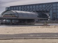 a building that is sitting on the beach by water with buildings in the background and people standing near it