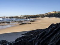 a sandy beach with blue water, rocks and cliffs in the background and waves breaking on the shore