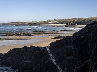 a sandy beach with blue water, rocks and cliffs in the background and waves breaking on the shore