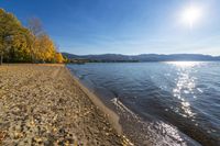 a sandy beach next to a lake under a bright sun filled sky in the distance