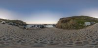 a fisheye photo of a sandy beach with stones and grass and the water in the background