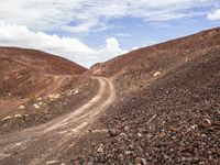 a gravel road leads up to a steep mountain side side with sparse rocks, a few white clouds and one pointy sky