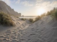 there is a trail on this sand dune near the ocean at sunset, leading to the beach with some grass and mountains in the distance