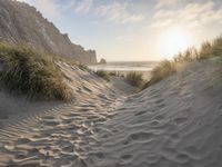 there is a trail on this sand dune near the ocean at sunset, leading to the beach with some grass and mountains in the distance