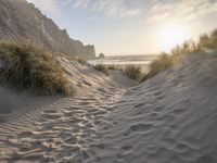 there is a trail on this sand dune near the ocean at sunset, leading to the beach with some grass and mountains in the distance