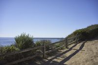 a fence and wooden rail on a dirt pathway next to the ocean beach and pier