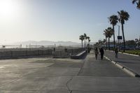 people walking along sidewalk next to walkway next to water area with palm trees and fenced in beach area