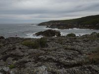 the man is standing near the ocean on the rocks next to the water, and one person is para sailing