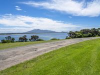 road by the water with a grassy and green field near the ocean and mountains in background