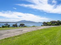 road by the water with a grassy and green field near the ocean and mountains in background