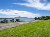 road by the water with a grassy and green field near the ocean and mountains in background