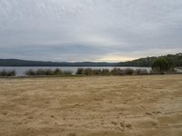 an empty sandy area at a lake in the woods with clouds overhead and on the ground to the right of the lake