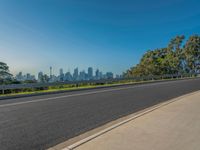 the road has been paved with black tarmac along it and is very wide, with trees lining side of it and a view of the city skyline