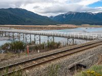 a small long rail track near water and mountains in the background that has an elevated bridge over it