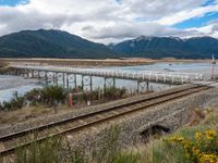 a small long rail track near water and mountains in the background that has an elevated bridge over it