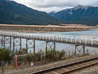 a small long rail track near water and mountains in the background that has an elevated bridge over it