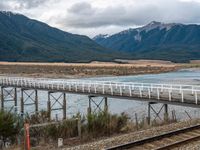 a small long rail track near water and mountains in the background that has an elevated bridge over it