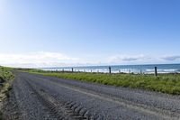 a gravel road that is surrounded by grass and water and the beach is in the background