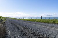 a gravel road that is surrounded by grass and water and the beach is in the background