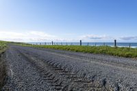 a gravel road that is surrounded by grass and water and the beach is in the background