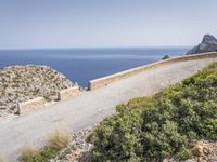 two motorcycles traveling on a narrow paved road by the ocean with mountains in the background