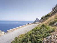 two motorcycles traveling on a narrow paved road by the ocean with mountains in the background