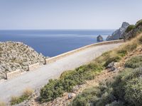 two motorcycles traveling on a narrow paved road by the ocean with mountains in the background