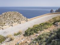 two motorcycles traveling on a narrow paved road by the ocean with mountains in the background