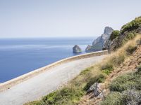 two motorcycles traveling on a narrow paved road by the ocean with mountains in the background