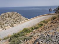 two motorcycles traveling on a narrow paved road by the ocean with mountains in the background