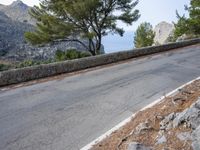 a paved road with some trees and a stone wall along the side of it and mountains in the background