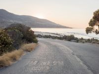the sun is setting behind some trees and an ocean line road with a man sitting on a motorcycle