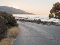 the sun is setting behind some trees and an ocean line road with a man sitting on a motorcycle