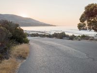the sun is setting behind some trees and an ocean line road with a man sitting on a motorcycle