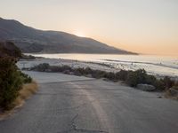 the sun is setting behind some trees and an ocean line road with a man sitting on a motorcycle