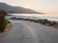 the sun is setting behind some trees and an ocean line road with a man sitting on a motorcycle