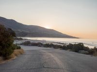 the sun is setting behind some trees and an ocean line road with a man sitting on a motorcycle