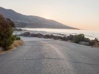 the sun is setting behind some trees and an ocean line road with a man sitting on a motorcycle
