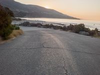 the sun is setting behind some trees and an ocean line road with a man sitting on a motorcycle