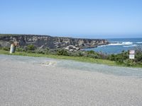 an empty road next to a cliff on a sunny day with ocean behind it and rocks, cliffs, bushes, grass