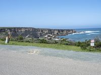 an empty road next to a cliff on a sunny day with ocean behind it and rocks, cliffs, bushes, grass