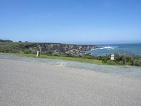 an empty road next to a cliff on a sunny day with ocean behind it and rocks, cliffs, bushes, grass