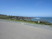 an empty road next to a cliff on a sunny day with ocean behind it and rocks, cliffs, bushes, grass