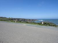 an empty road next to a cliff on a sunny day with ocean behind it and rocks, cliffs, bushes, grass